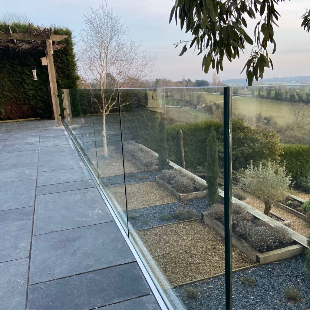 A glass railing separates a tiled patio from a garden with raised beds, gravel paths, and various trees, overlooking a scenic rural landscape with fields and distant trees.