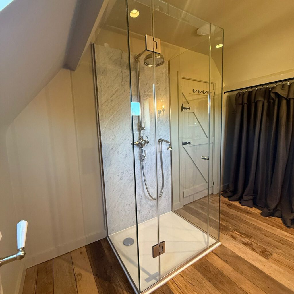 Glass-enclosed shower with a marble wall and rainfall showerhead in a bathroom with wooden floors, white paneled walls, and a black curtain along one side.