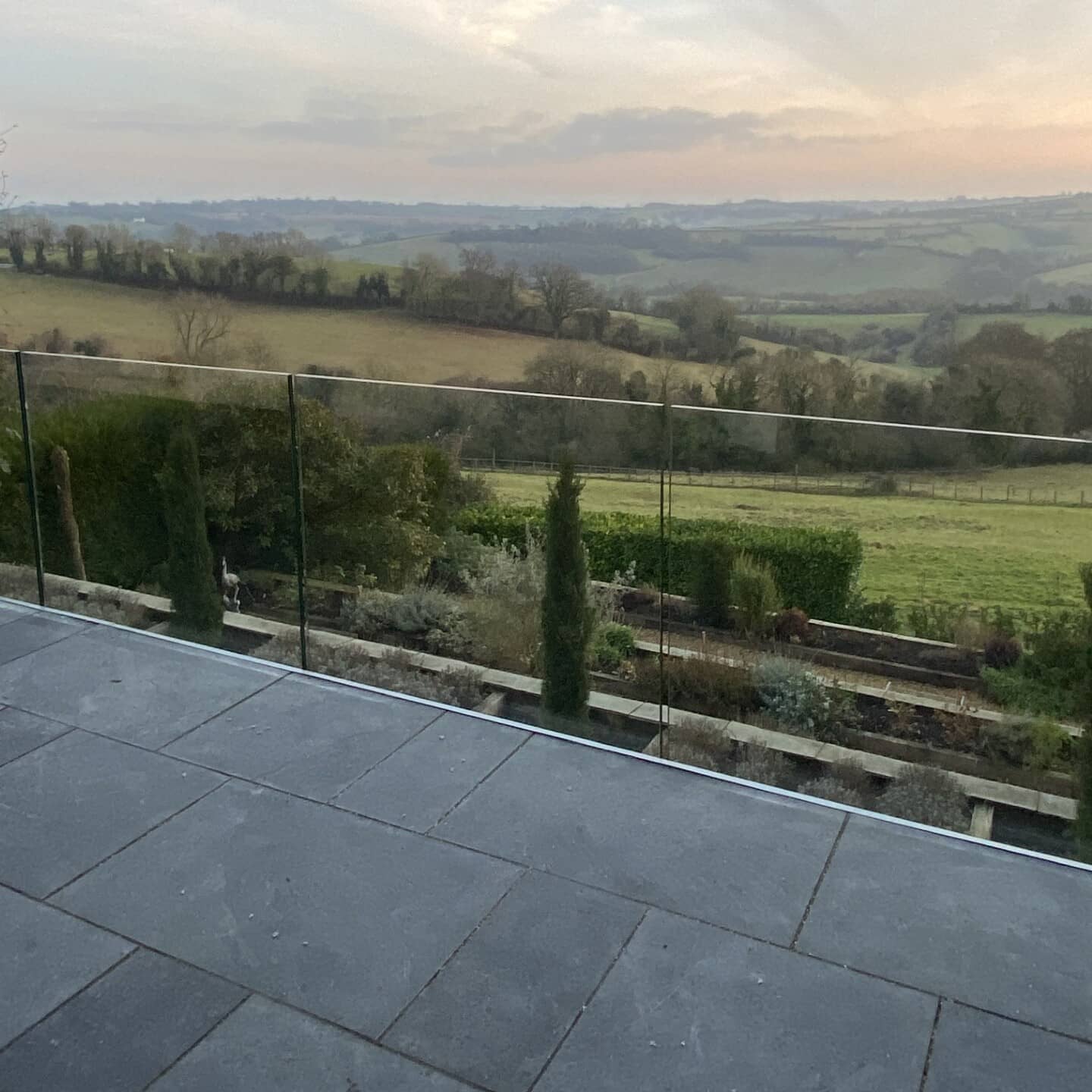 A view from a stone-tiled patio with a glass railing overlooking a green, rolling countryside landscape at sunset, with fields, hedges, and distant hills under a cloudy sky.