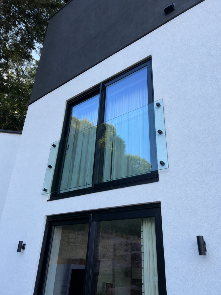 A modern house facade features large windows with a clear glass balcony railing, white walls, and a black upper section. Outdoor wall lights are mounted beside the lower window. Trees are reflected in the upper window.