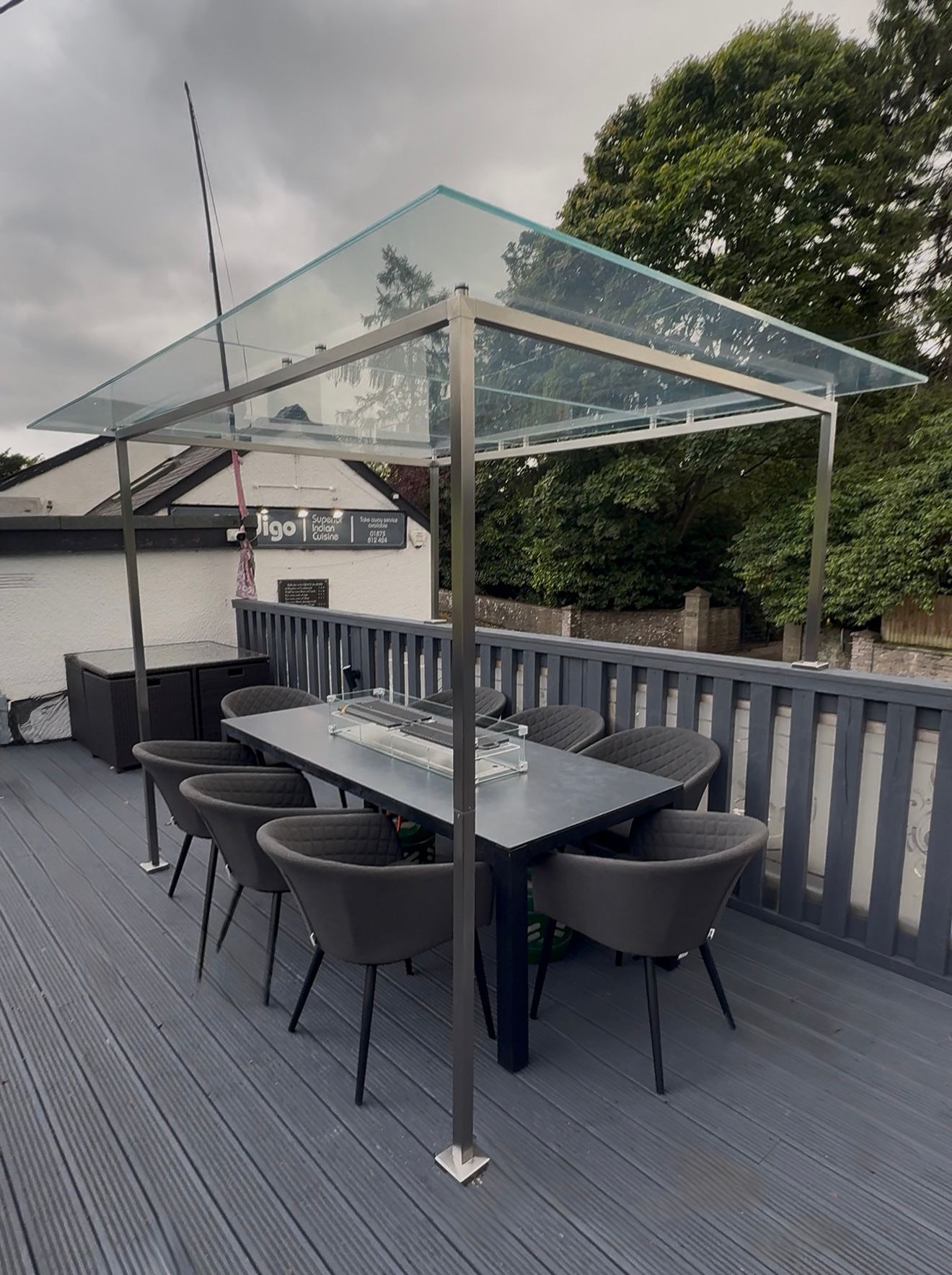 A modern outdoor dining area on a gray deck features a rectangular table with eight dark chairs and a transparent glass roof canopy supported by metal poles. Trees and a white building are visible in the background.