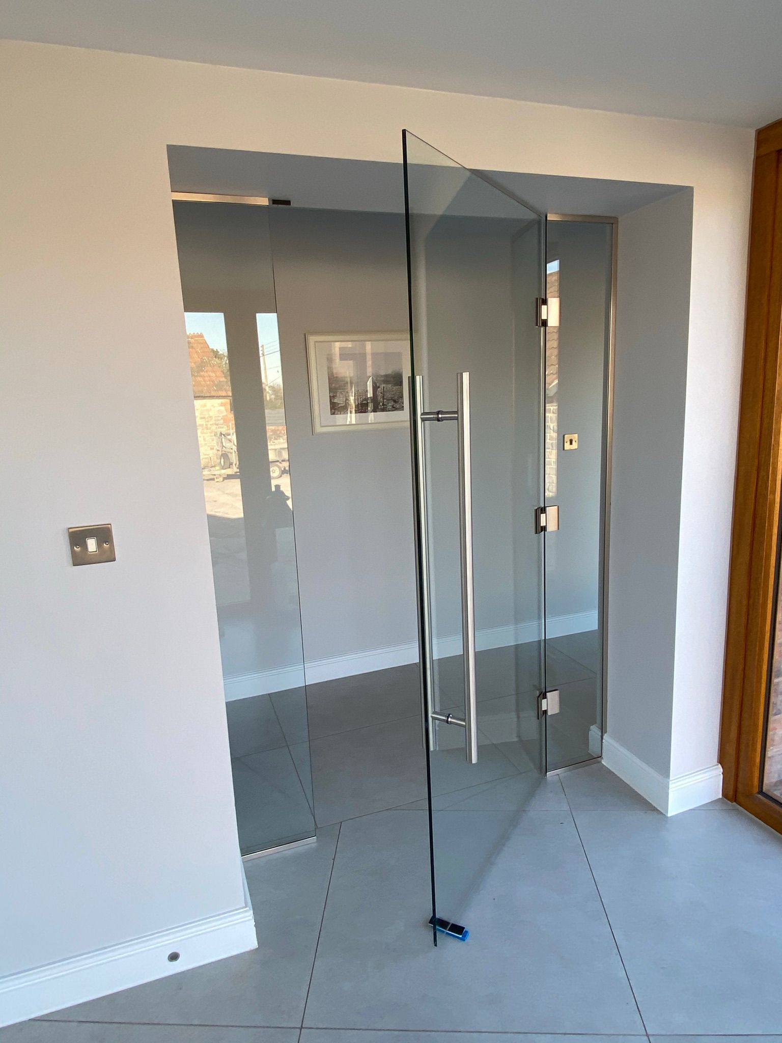 A frameless glass door is partially open in a modern, minimalistic room with light-colored walls and tiled floors. There is a photo frame on the far wall and sunlight coming through nearby windows.