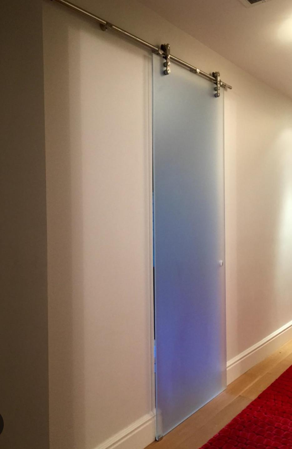 A frosted glass sliding door with a metal rail is installed in a white hallway with light wood flooring and a red textured rug on the floor.