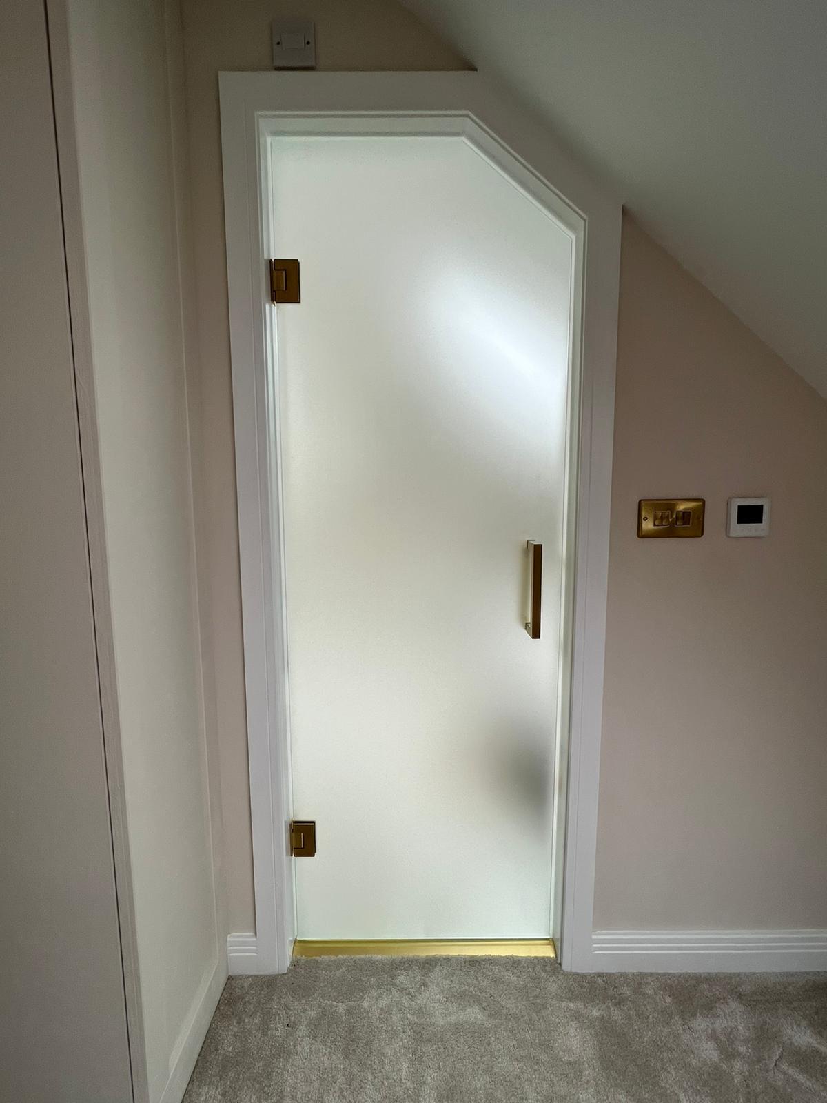 A frosted glass door with a white frame and brass hardware is set under a sloped ceiling. The door is next to a light switch and a thermostat on a cream-colored wall above a light gray carpet.