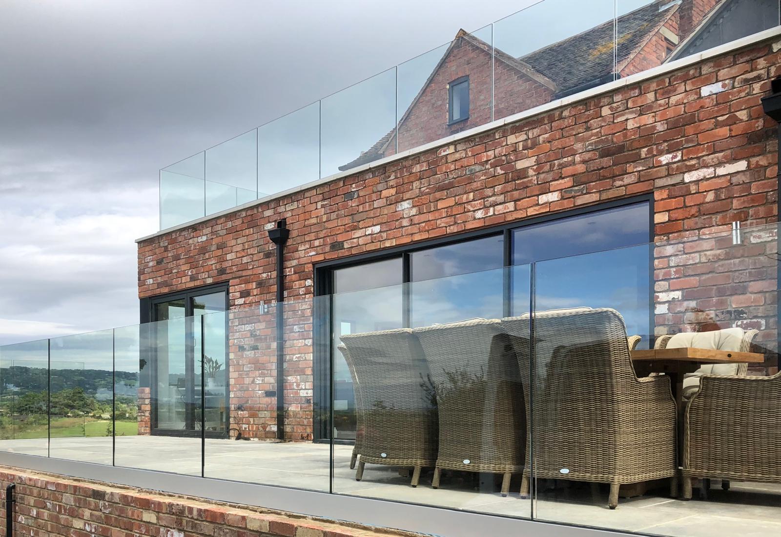 A modern patio with wicker chairs and a wooden table sits behind glass railings on the upper floor of a brick building, with large sliding glass doors and countryside visible in the background.