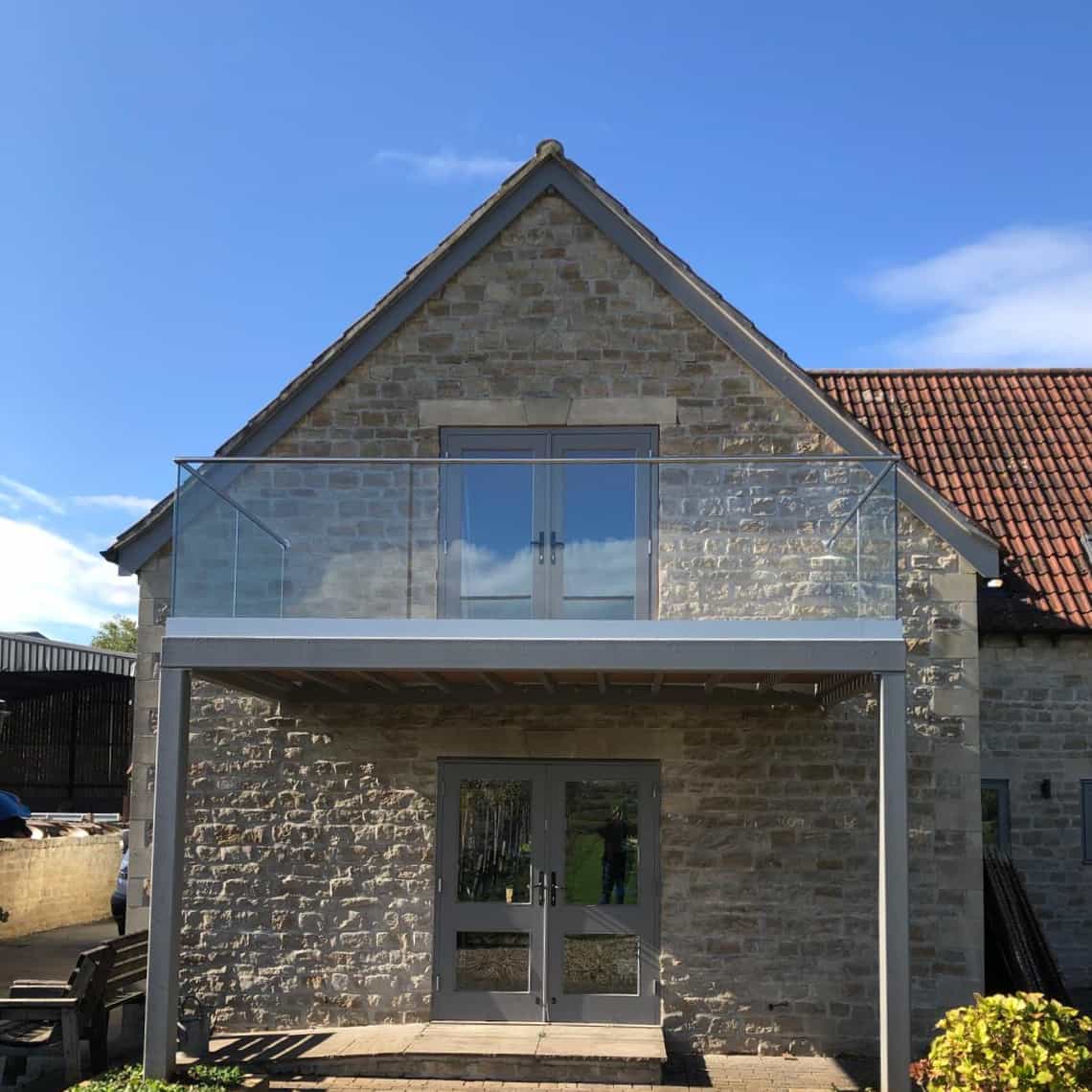A stone house with a glass balcony and double doors on both the ground floor and upper floor, under a clear blue sky. The balcony has a modern glass railing and metal frame.