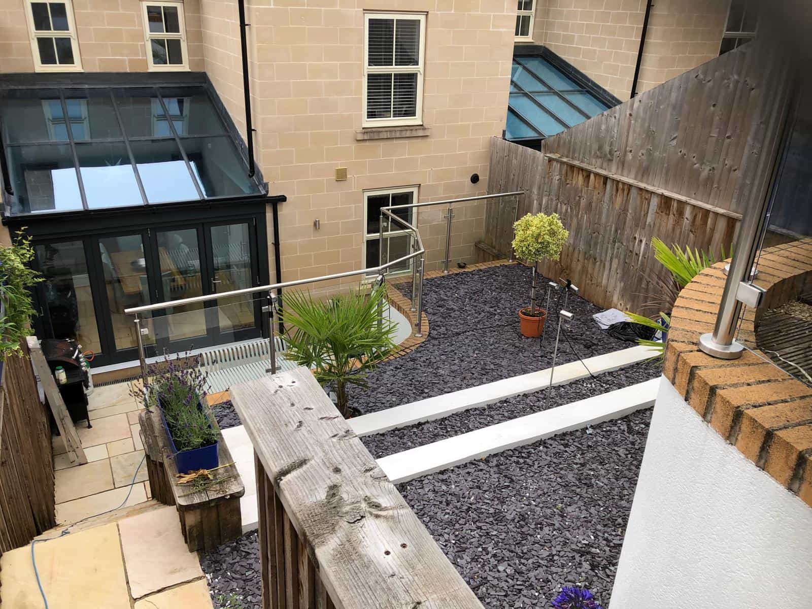 Modern backyard with slate chippings, potted plants, glass railings, and a patio area. Steps lead down to a glass conservatory attached to a beige brick building with large windows.