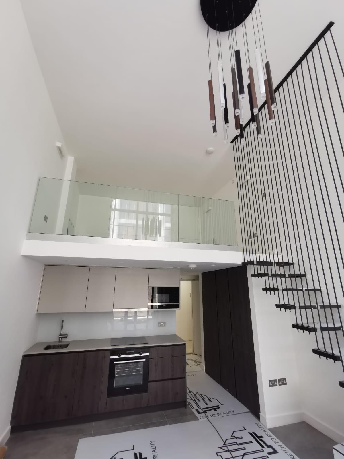 Modern loft apartment with a minimalist kitchen, dark cabinets, built-in appliances, and a mezzanine level with glass railing. A staircase with vertical railings and a contemporary hanging light fixture complete the space.