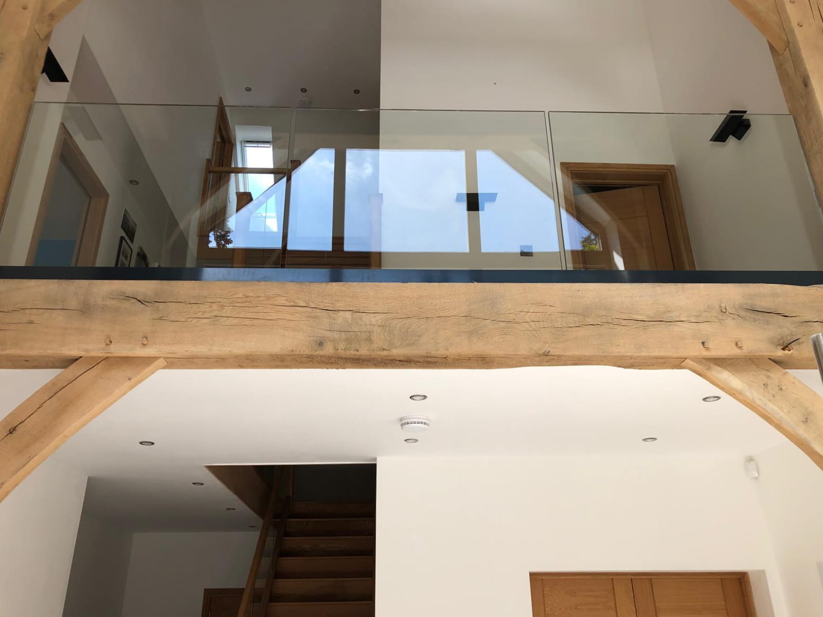 View of a modern interior with exposed wooden beams, a glass balcony railing, staircase, and wooden doors. The space is bright with natural light and white walls.