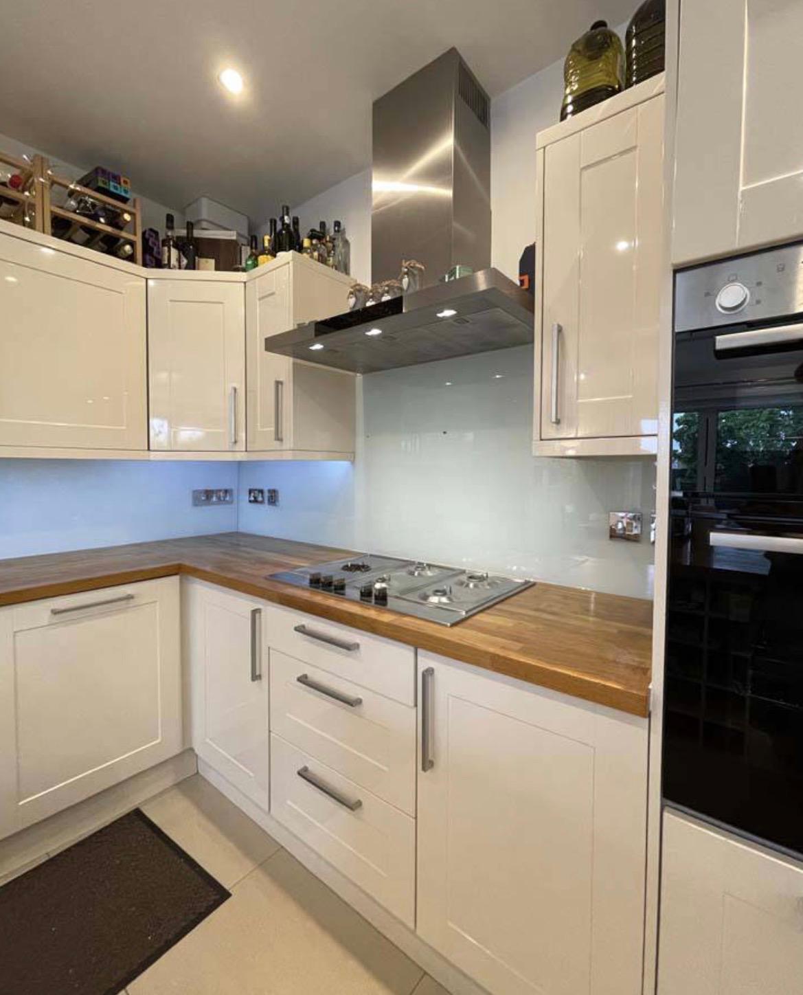 Modern kitchen with cream cabinets, wooden countertops, a built-in gas cooktop, stainless steel oven, and extractor hood. Shelves with bottles are above the cabinets, and a window reflects light on the right.