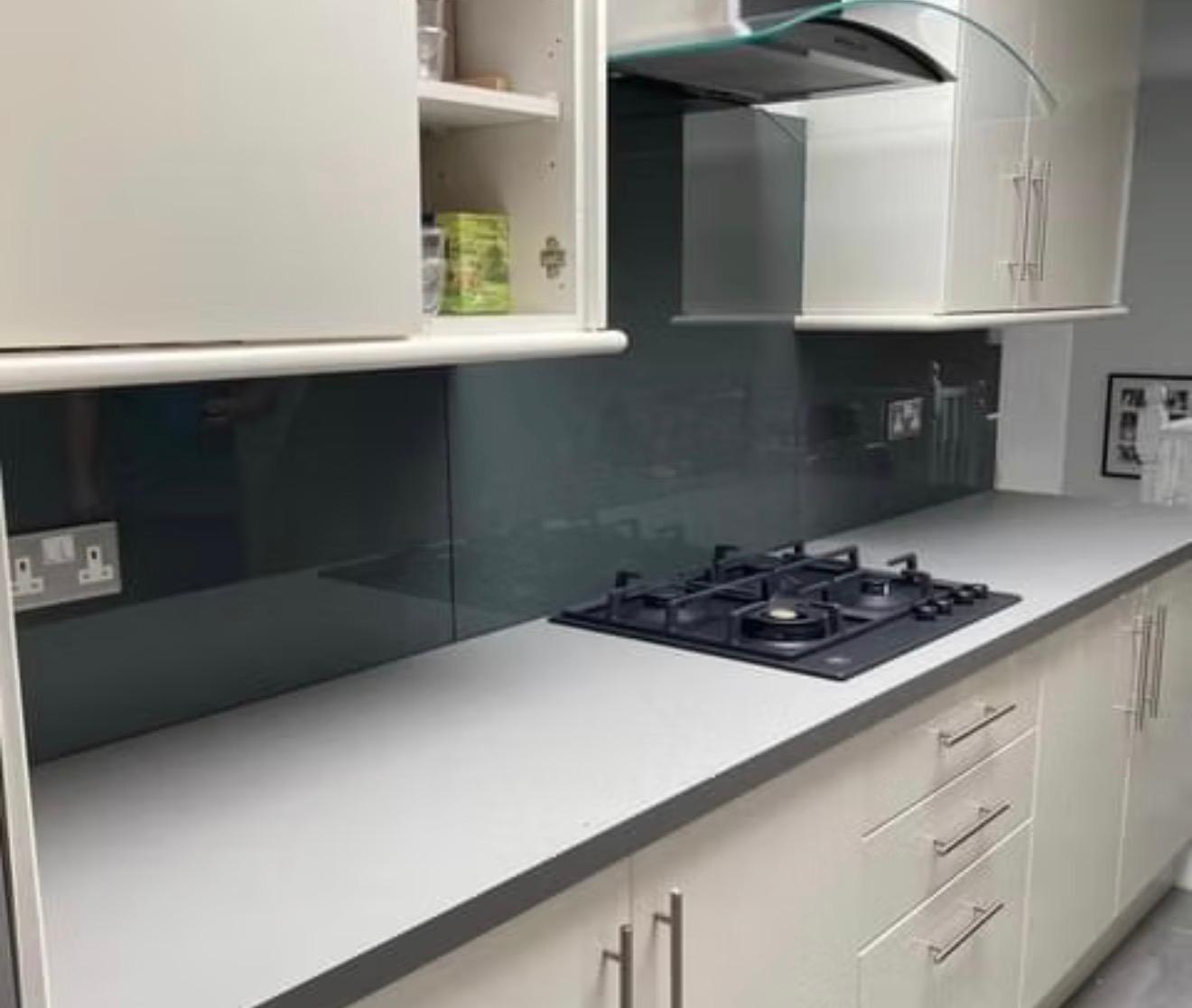 Modern kitchen with white cabinets and drawers, a grey countertop, a built-in black gas cooktop, glass backsplash, and overhead extractor hood. Some shelves are open, displaying pantry items.