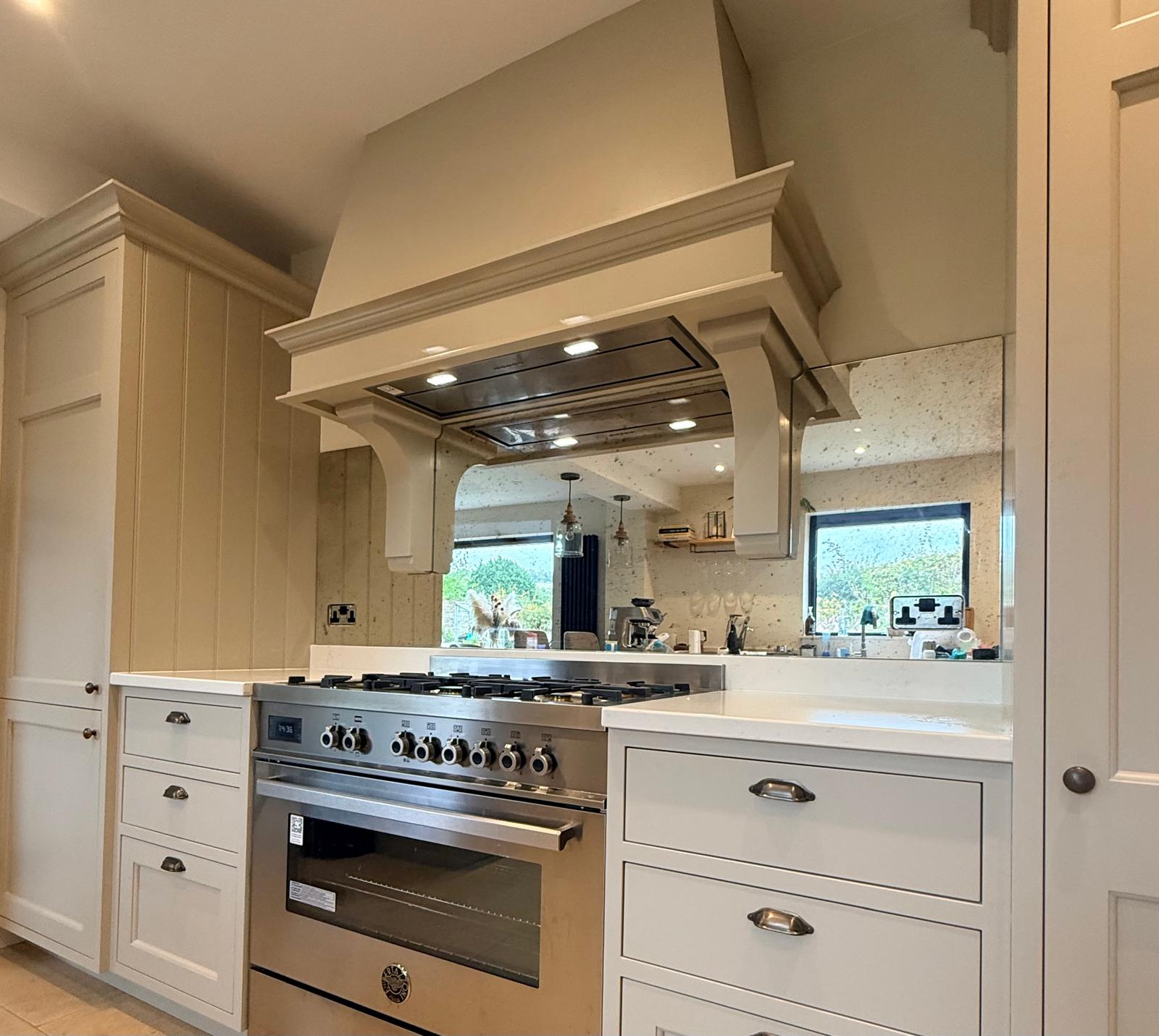 Modern kitchen featuring a large stainless steel gas stove with multiple burners, white cabinetry, a range hood, and a mirrored backsplash reflecting the rest of the kitchen and outdoor scenery.