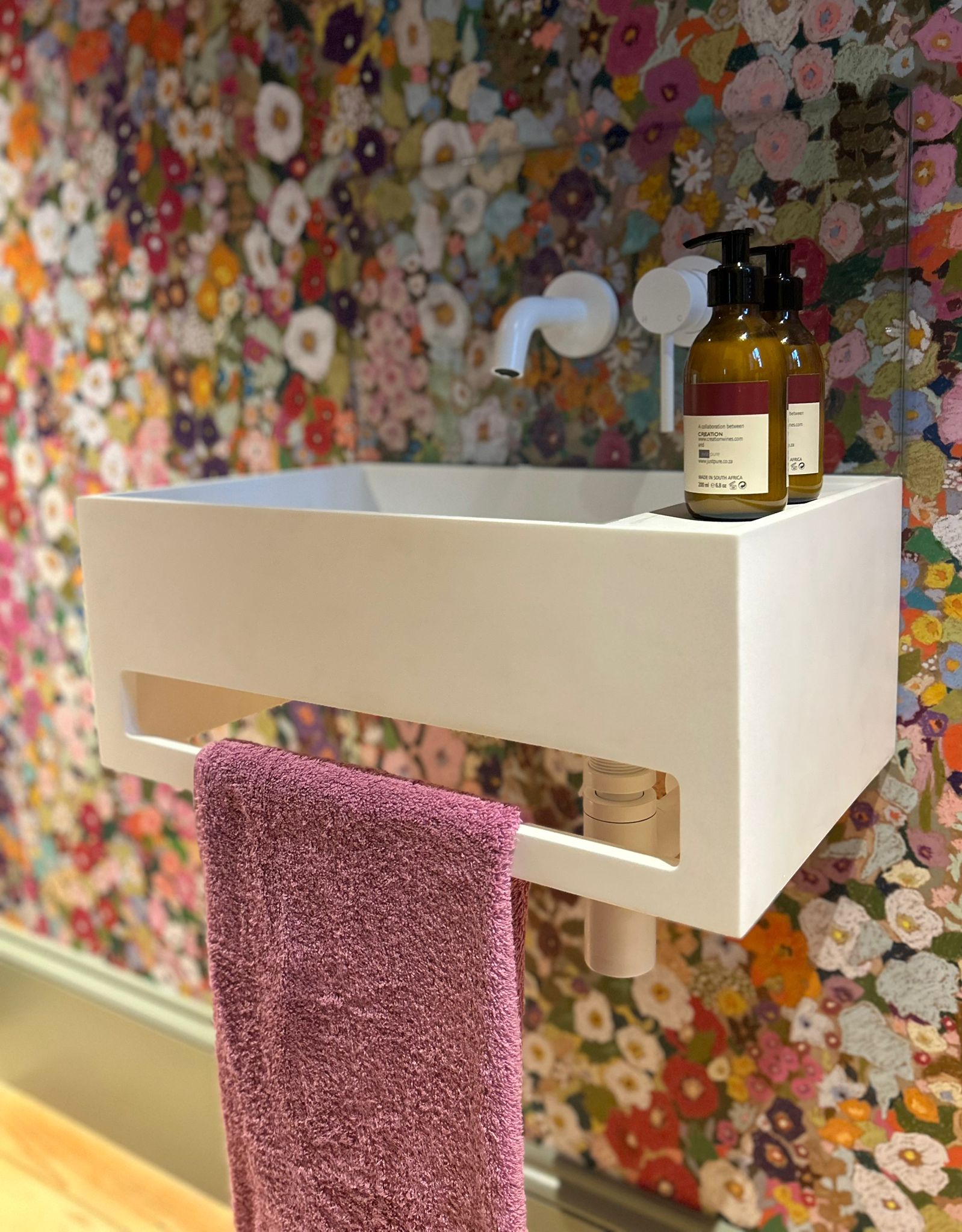 A modern white wall-mounted sink with a purple towel hanging below and two brown soap bottles on top, set against a colorful floral wallpaper background.