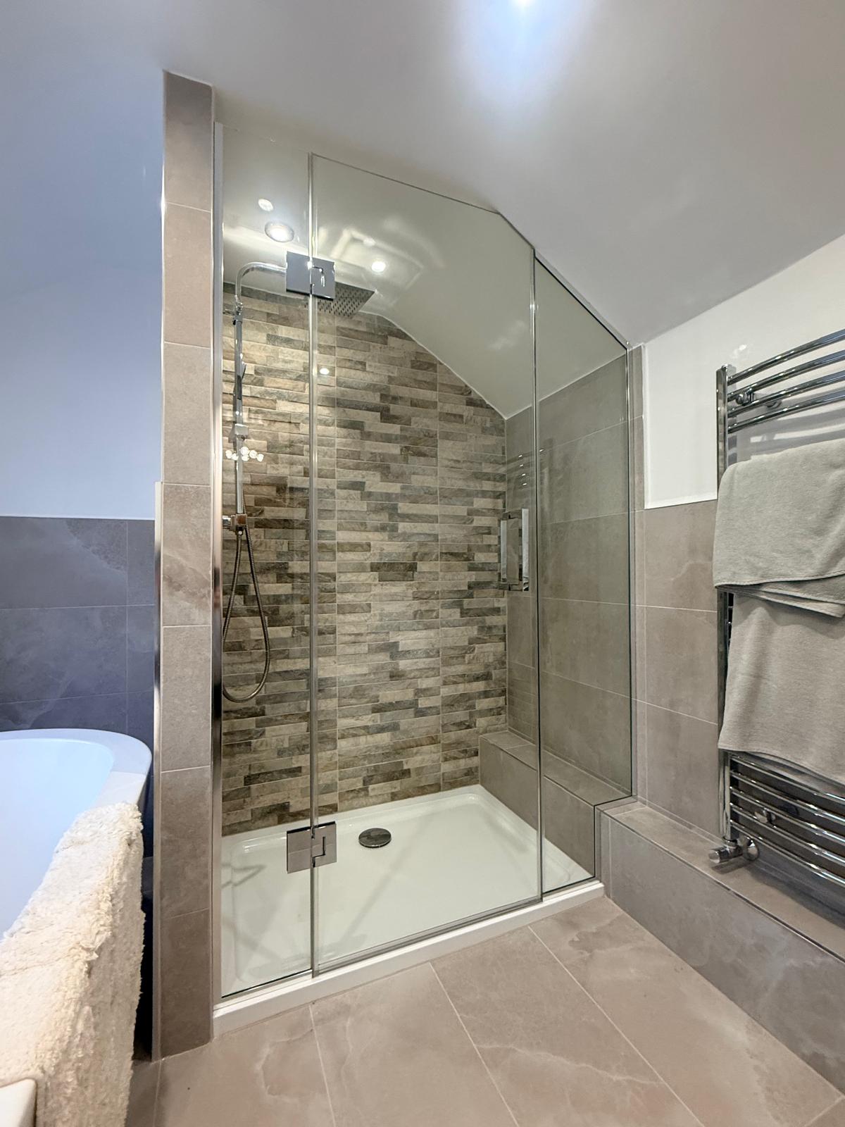 Modern bathroom with a glass-enclosed shower featuring stone-patterned tiles, a rainfall showerhead, and a handheld sprayer. A heated towel rail with towels is on the right, and part of a bathtub is visible on the left.