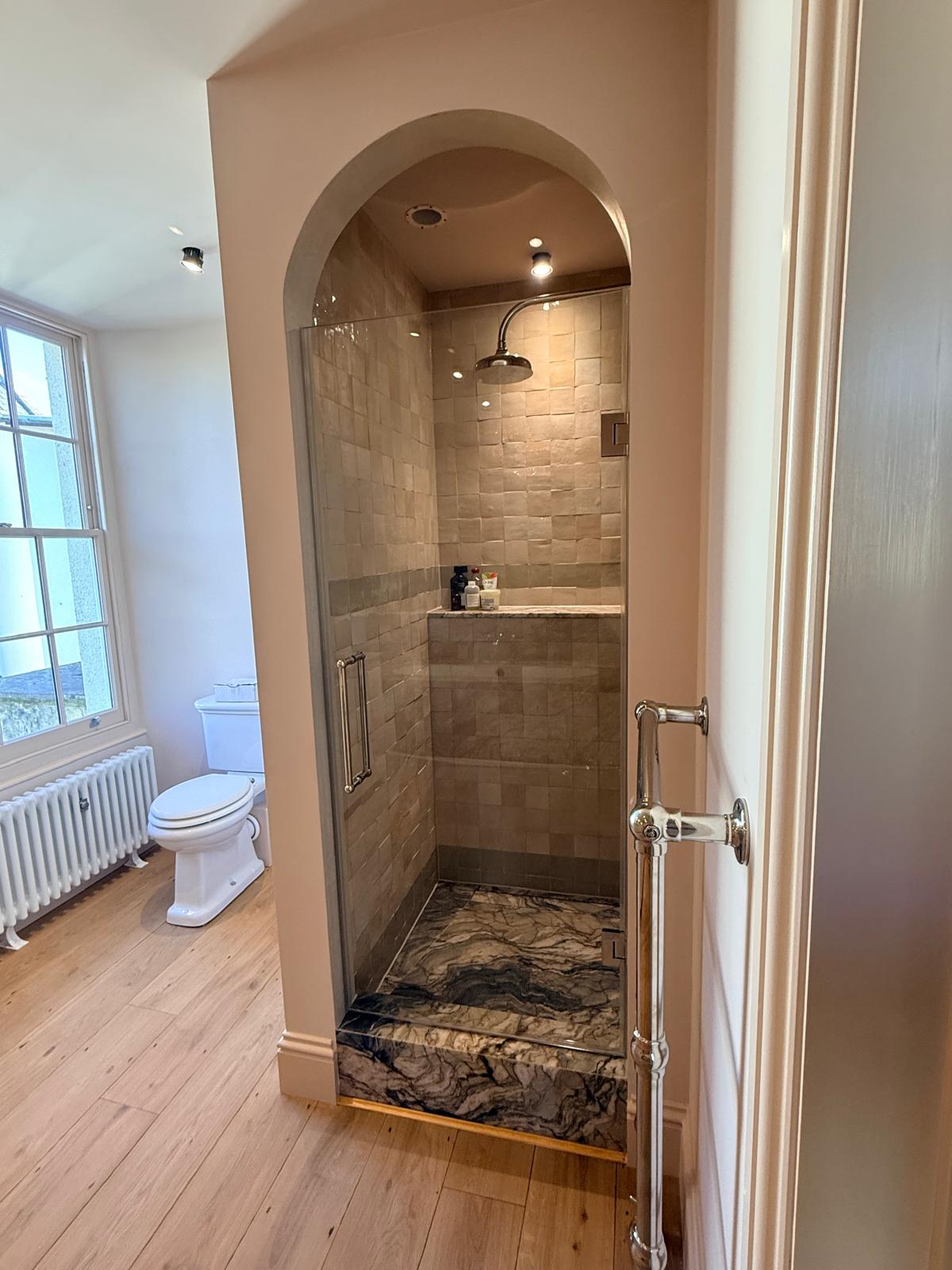 A bathroom with a glass-enclosed shower featuring beige tiles and a marble base, next to a toilet and a large window with white trim. The floor is light wood, and a chrome towel rail is mounted on the wall.