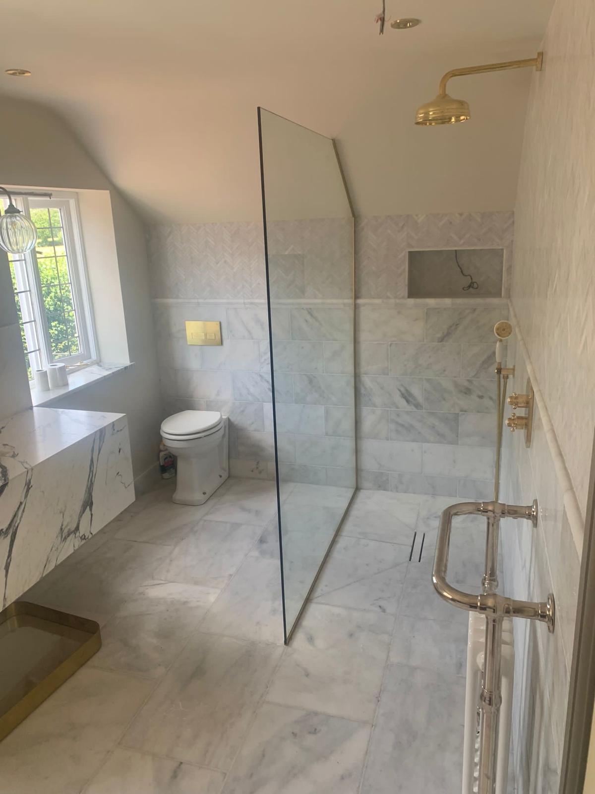 A modern bathroom with marble tiles, a walk-in shower with a glass divider, a gold showerhead, a floating marble vanity, a wall-mounted toilet, and a large window letting in natural light.