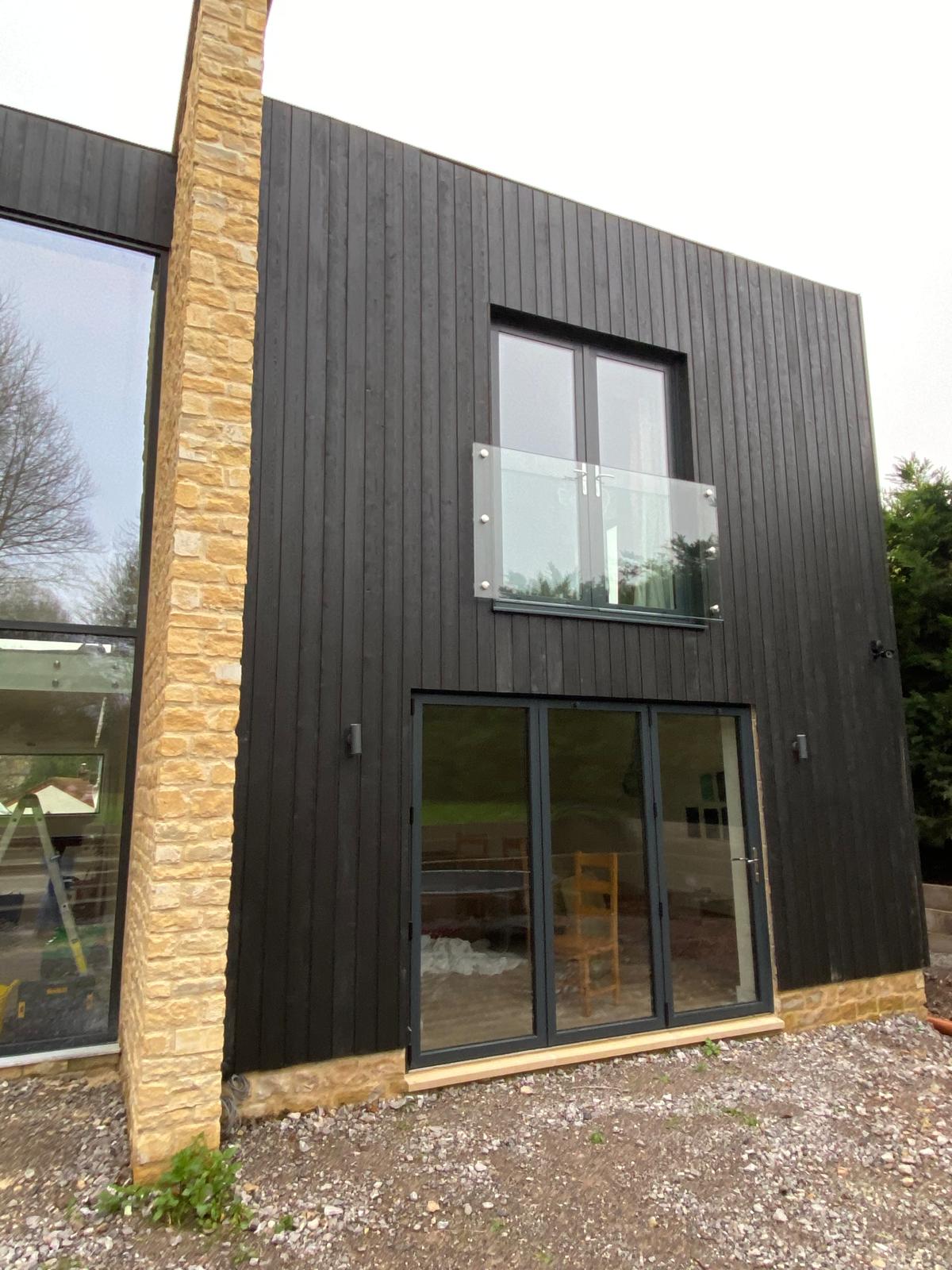 A modern two-story building with black vertical wooden cladding, stone accents, large glass doors on the ground floor, and a glass balcony railing on the upper floor.