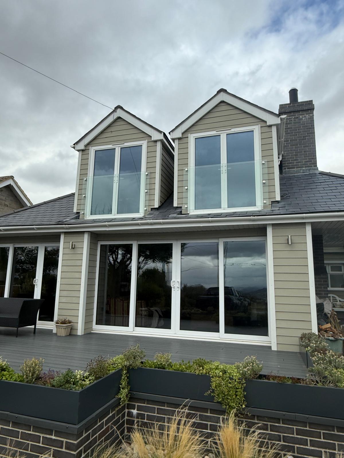 A modern house with light gray horizontal siding, two large dormer windows upstairs, and wide glass sliding doors on the ground floor. There are planter boxes and a small garden in front of a raised deck.
