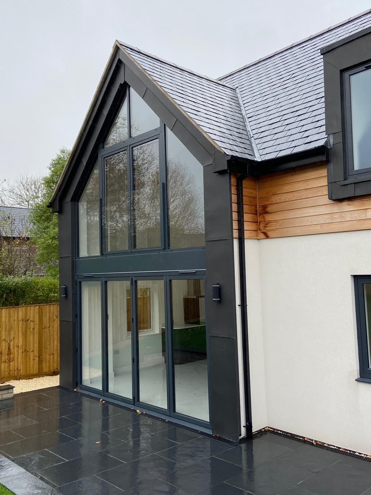 Modern house with large floor-to-ceiling glass windows, slate roof, light wood and white exterior walls, and a black tiled patio. There's a wooden fence and greenery visible in the background.