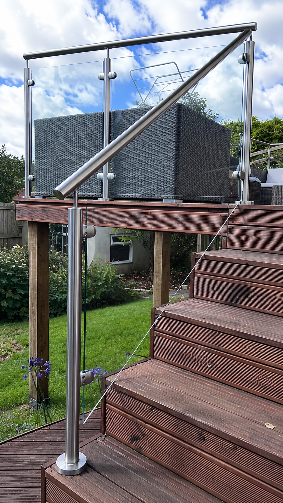 Wooden outdoor stairs with a modern metal and glass railing lead up to a raised deck. On the deck is a wicker furniture set. The area is surrounded by grass, plants, and trees under a partly cloudy sky.