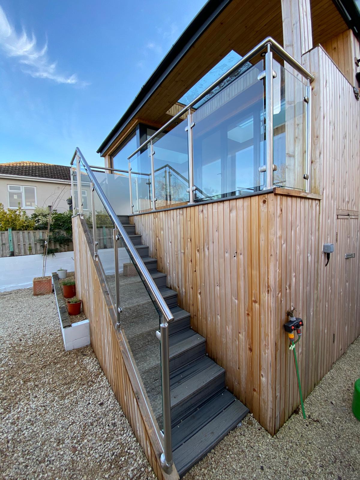 A modern wooden exterior staircase with metal handrails and glass balustrades leads up to a raised patio area attached to a wooden building, with potted plants and a hose nearby.