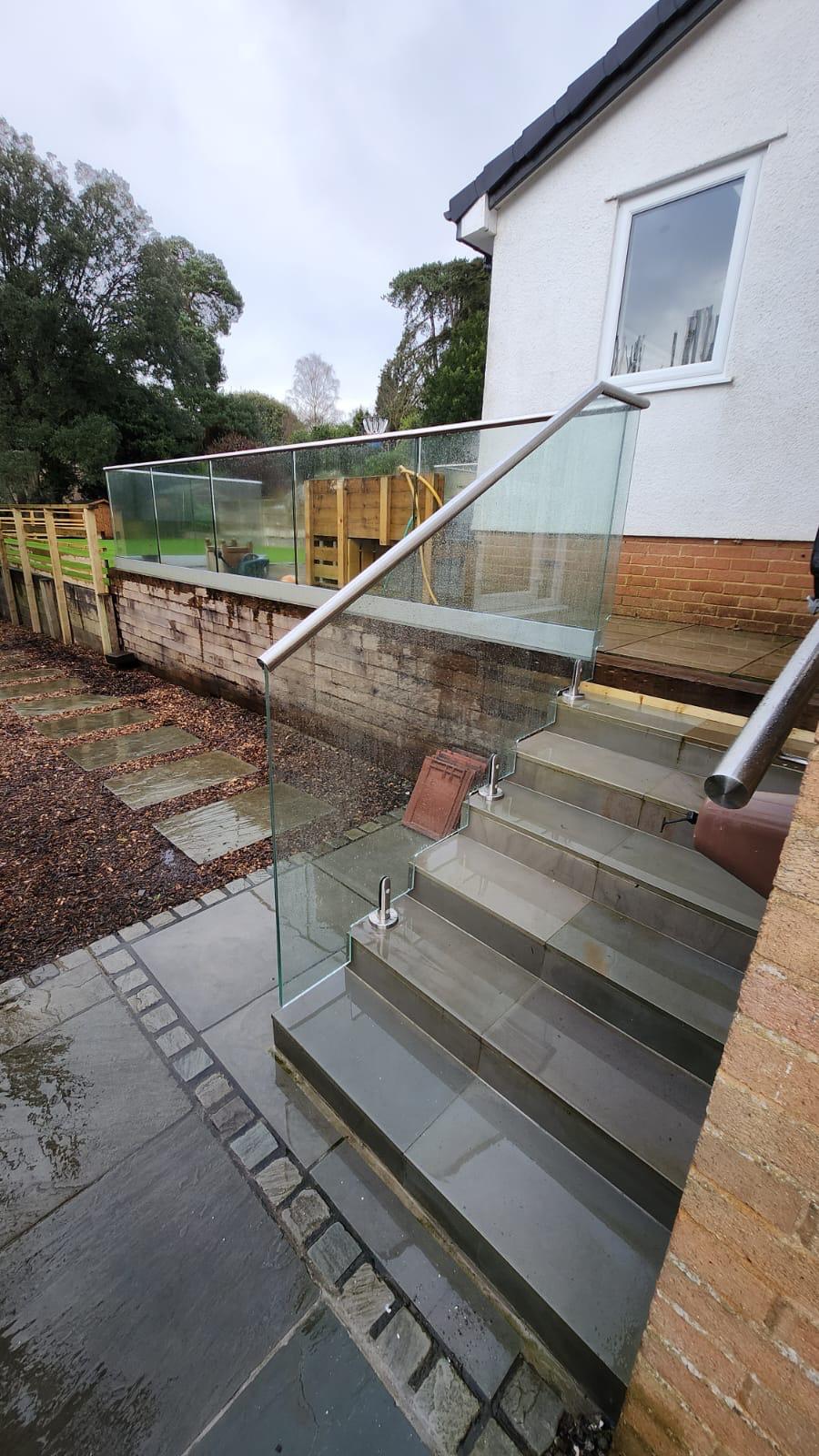 Glass balustrade with metal handrail lining outdoor stairs by a white house, leading down to a garden area with stone slabs and a lawn, enclosed by fences.