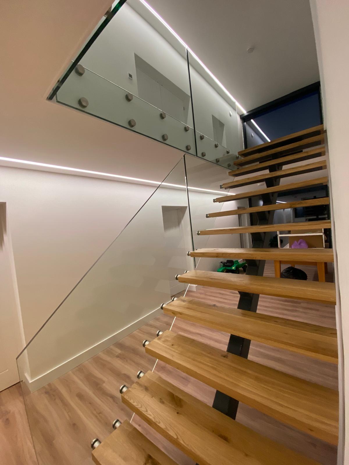 Modern wooden staircase with open risers and glass railing, leading to an upper floor with glass walls. The space has light-colored walls, wood flooring, and sleek, minimalist design elements.