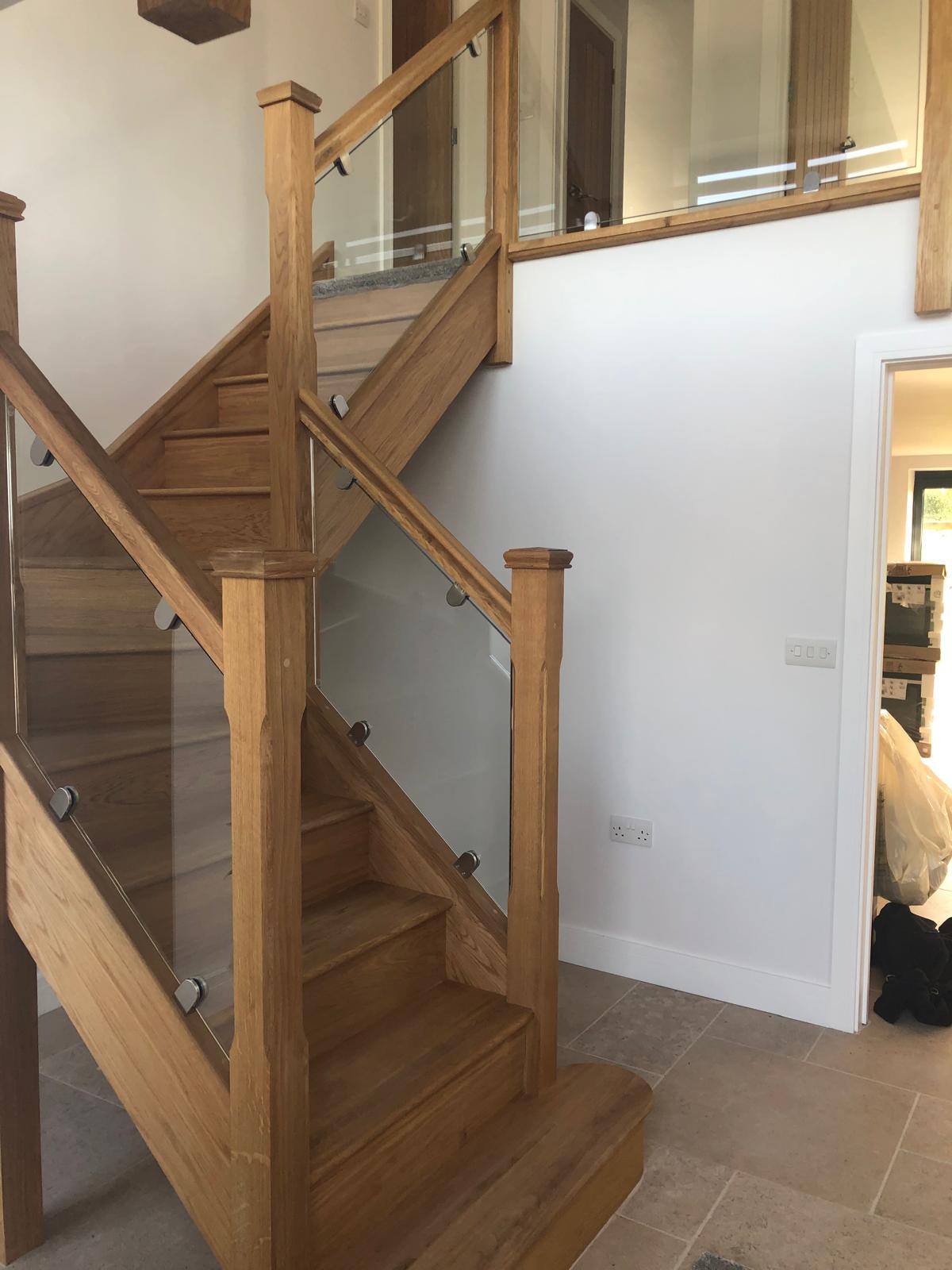 Modern wooden staircase with glass panel railings in a bright interior with white walls and beige tiled floor. An open doorway leads to another room with a window and some items visible inside.
