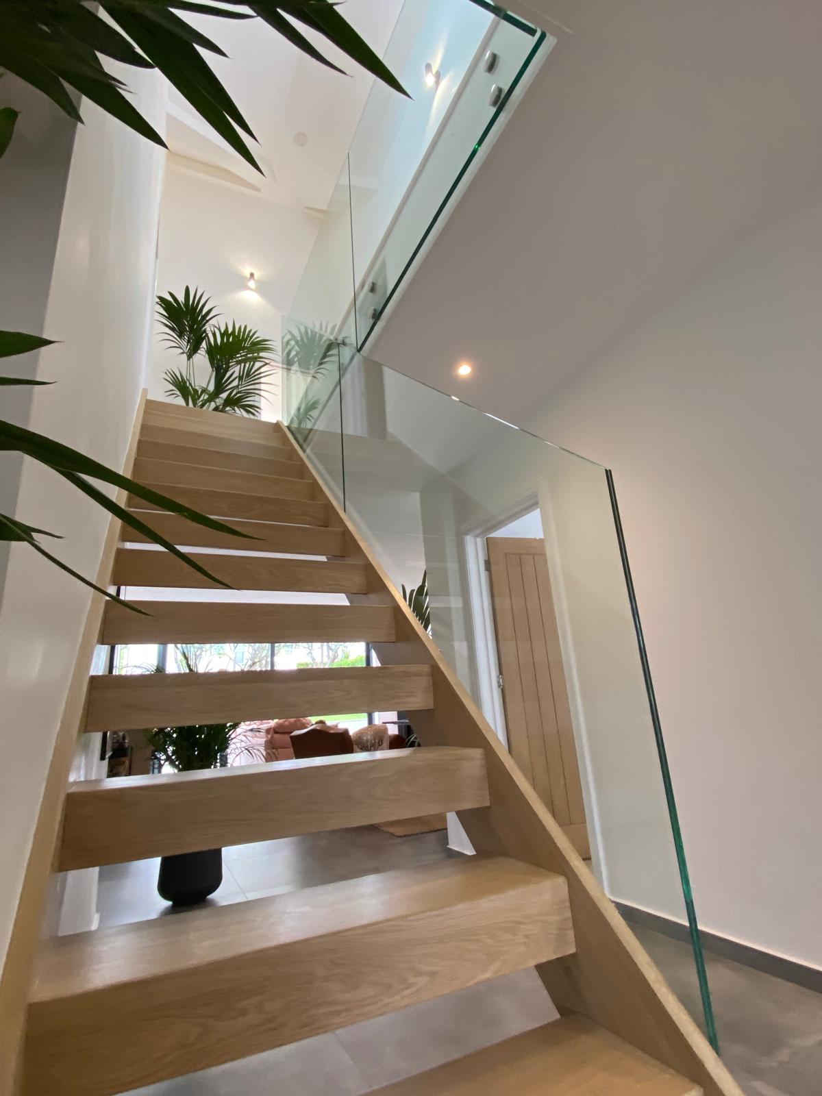 A modern wooden staircase with glass railings leads to an upper floor in a bright home. Potted plants decorate the space, and natural light streams in through the windows.
