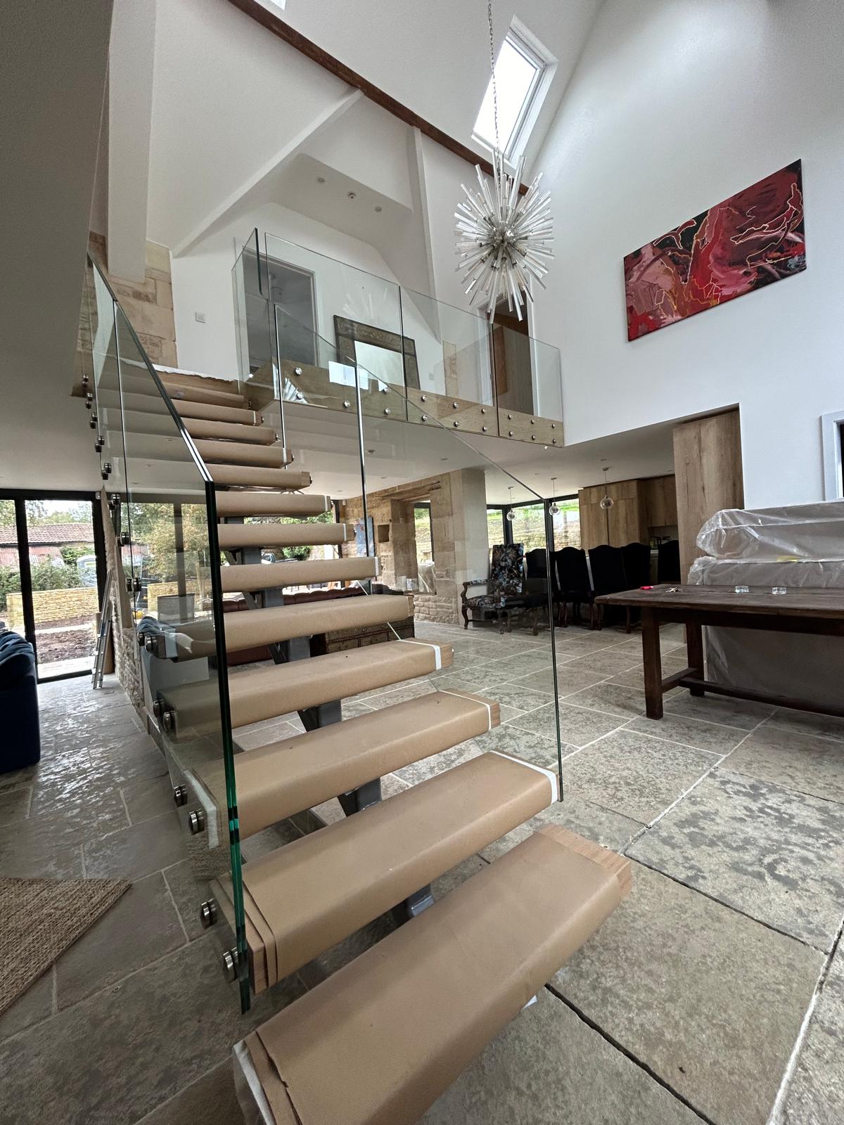 A modern living space with stone tile flooring, a glass-paneled staircase with covered steps, high ceilings, a dramatic light fixture, and a loft area. Large windows provide natural light; abstract art hangs on the wall.