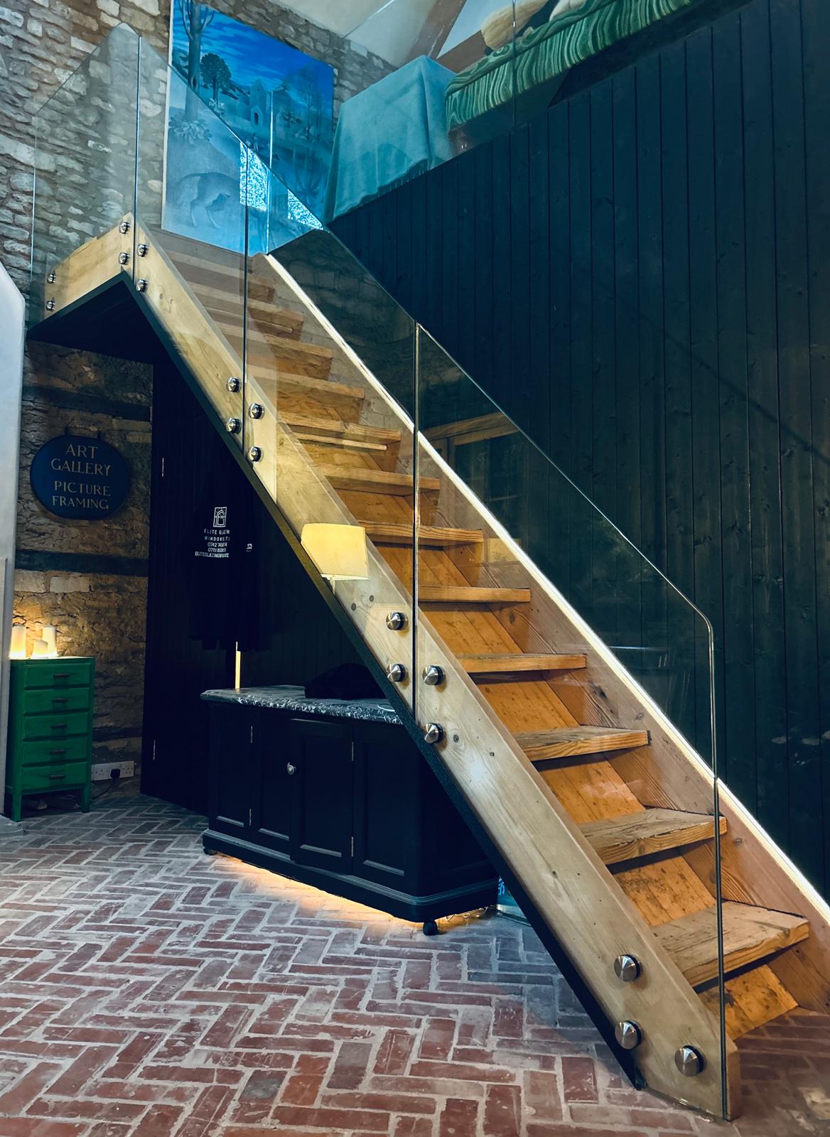 Modern wooden staircase with glass railings against a dark wall, set above a black cabinet on a red brick floor in a room with exposed stone walls and art gallery signage.