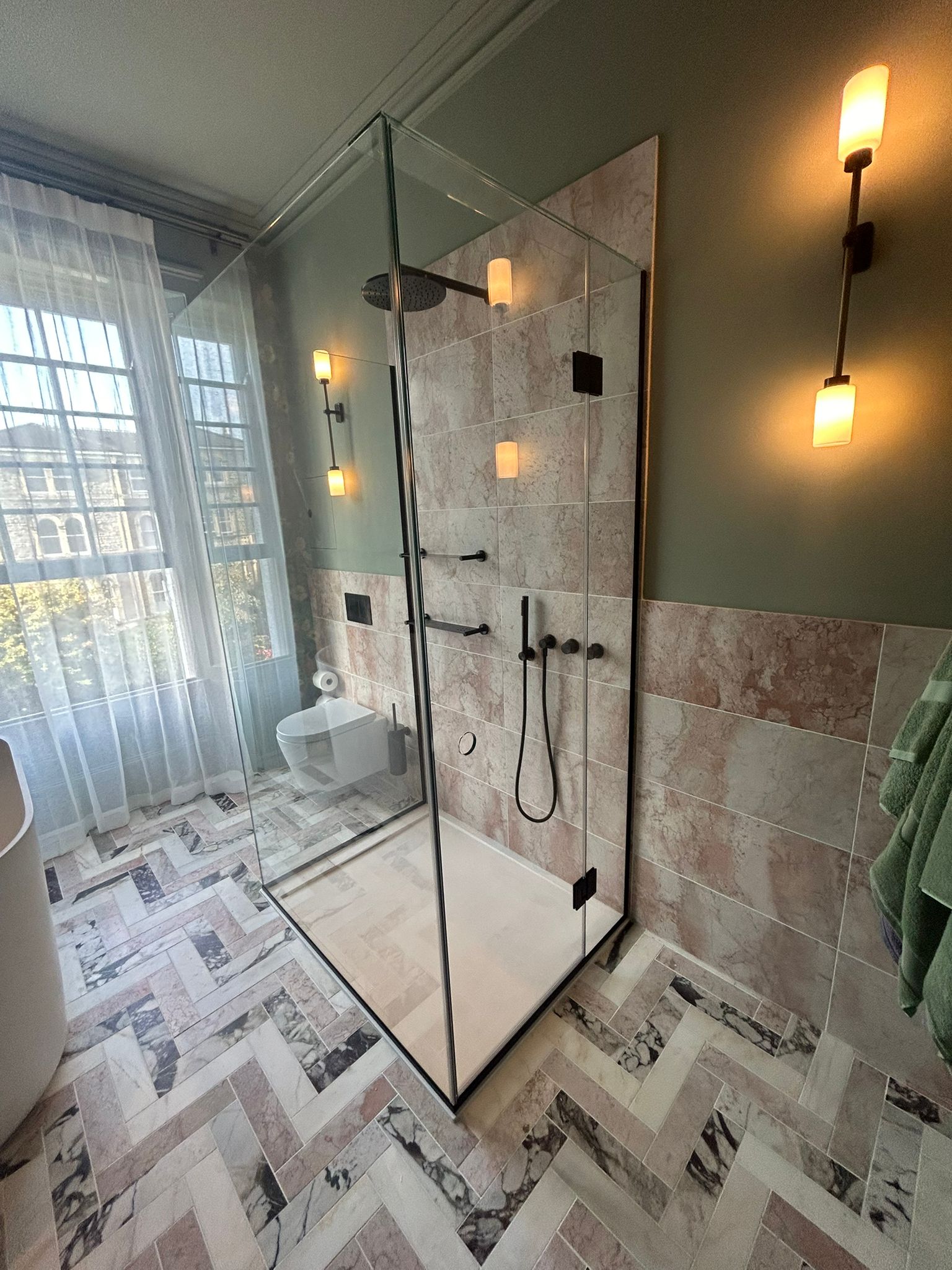 Modern bathroom with a glass shower enclosure, pink and gray marble tiles, a wall-mounted toilet, double wall lights, a towel hanging, and a large window with sheer white curtains letting in natural light.