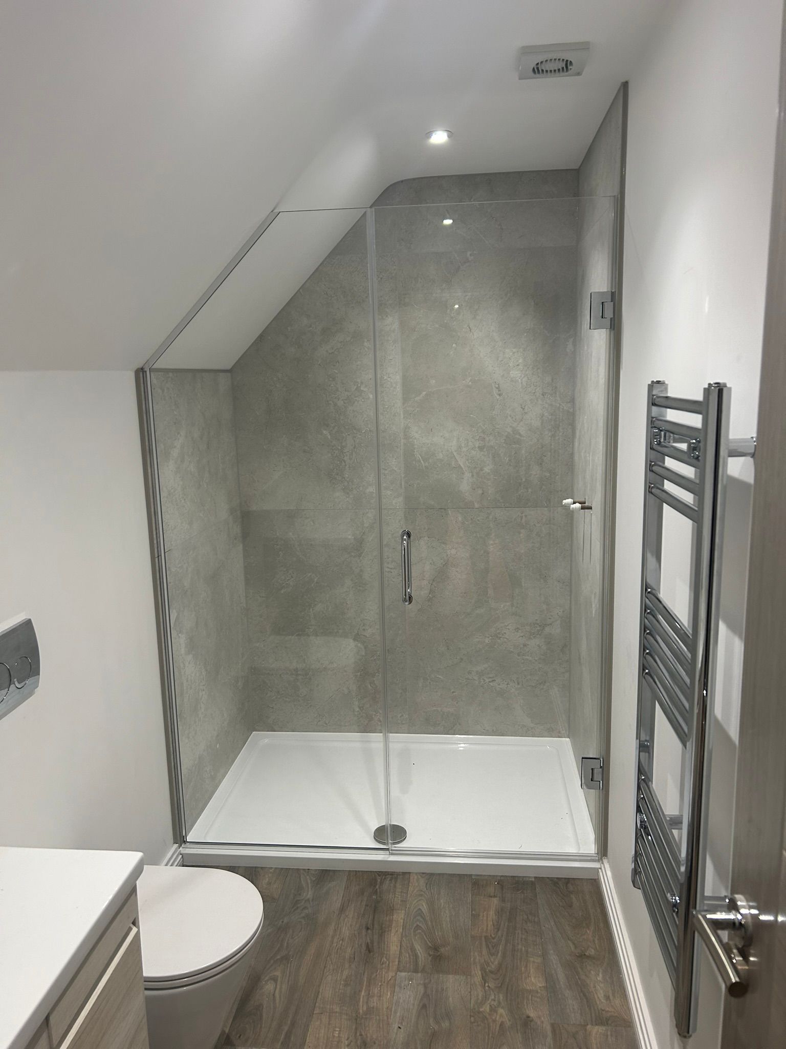 Modern bathroom with a glass-enclosed shower featuring gray tiled walls, a rainfall showerhead, a wall-mounted toilet, a heated towel rail, and wood-effect flooring under a sloped ceiling.