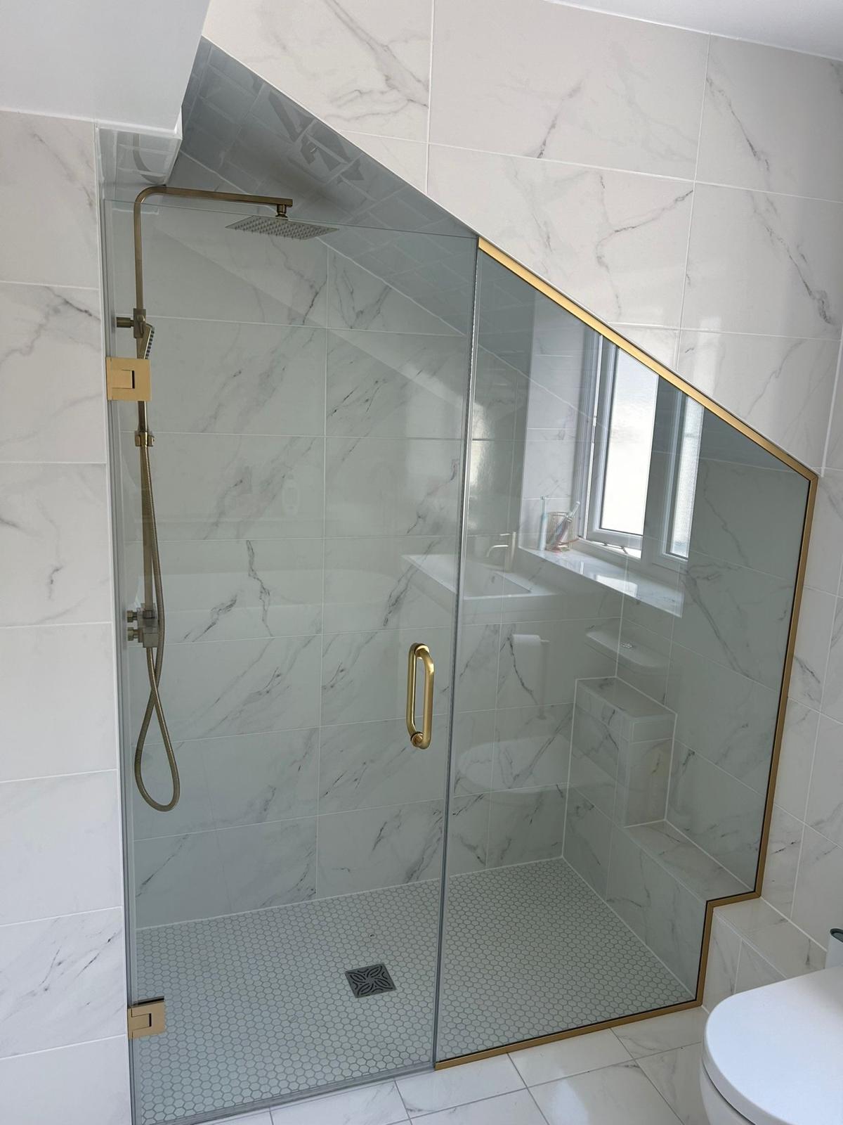 A modern shower with a glass enclosure featuring a gold trim, set against white marble-style tiles. The glass is cut diagonally to fit under a sloped ceiling, and there is a window next to the shower.