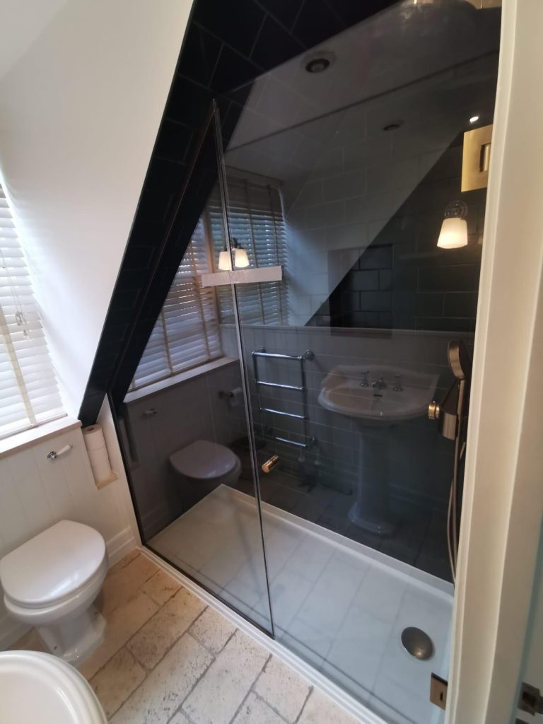 A modern bathroom with an angled ceiling, glass shower enclosure, white pedestal sink, wall-mounted towel warmer, toilet, and a window with blinds letting in natural light.