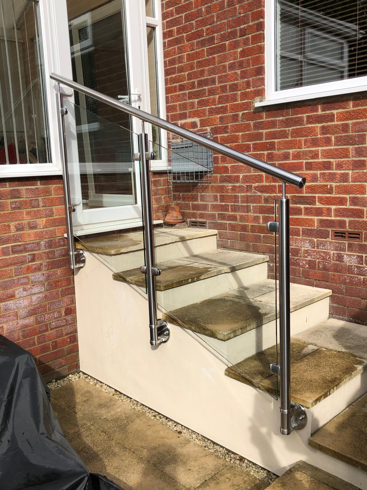 A small outdoor staircase with four stone steps and a modern metal handrail, leading to a white door set in a red brick wall. The handrail is shiny and mounted with metal brackets.