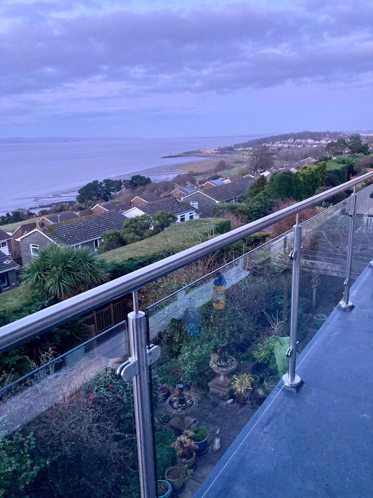 View from a balcony with glass railing overlooking houses, a garden with potted plants, and a coastline with the sea and cloudy sky in the distance.