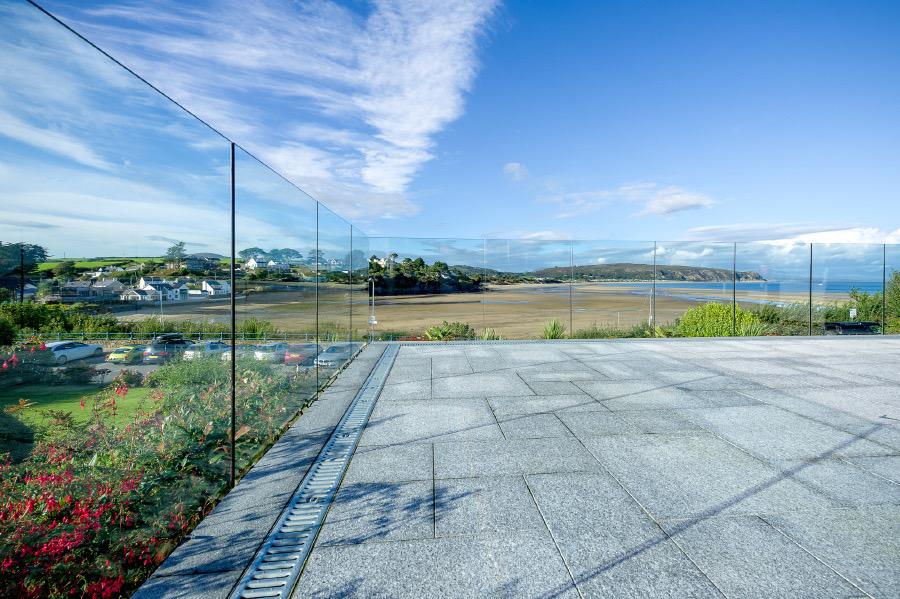 A glass railing encloses a stone patio overlooking a scenic coastal landscape with sandy shore, blue sea, parked cars, greenery, and a partly cloudy sky.
