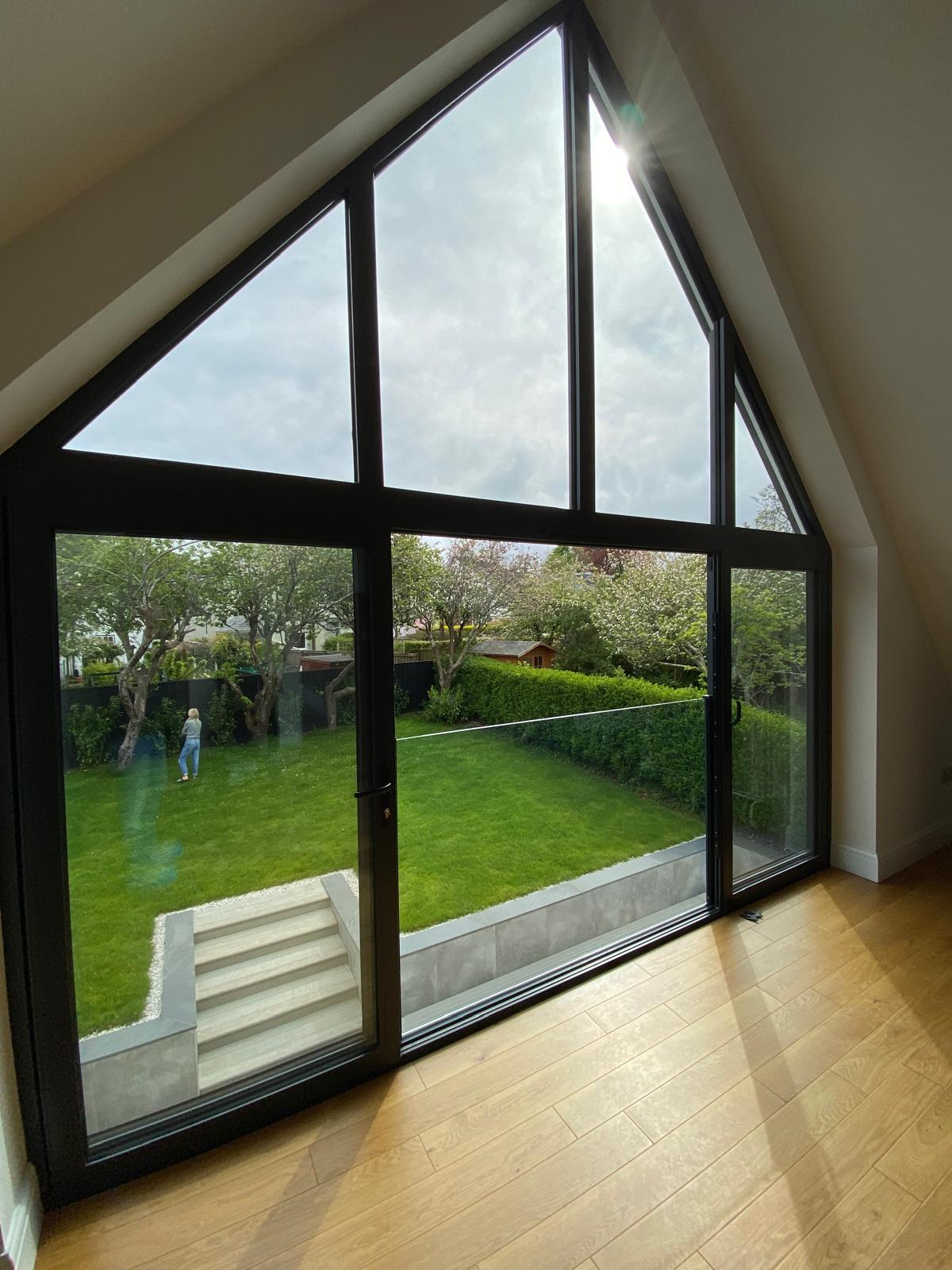 A large triangular window overlooks a green garden with trees, grass, and a small shed. Sunlight streams in, illuminating the wooden floor inside. A person stands in the garden near the hedge.
