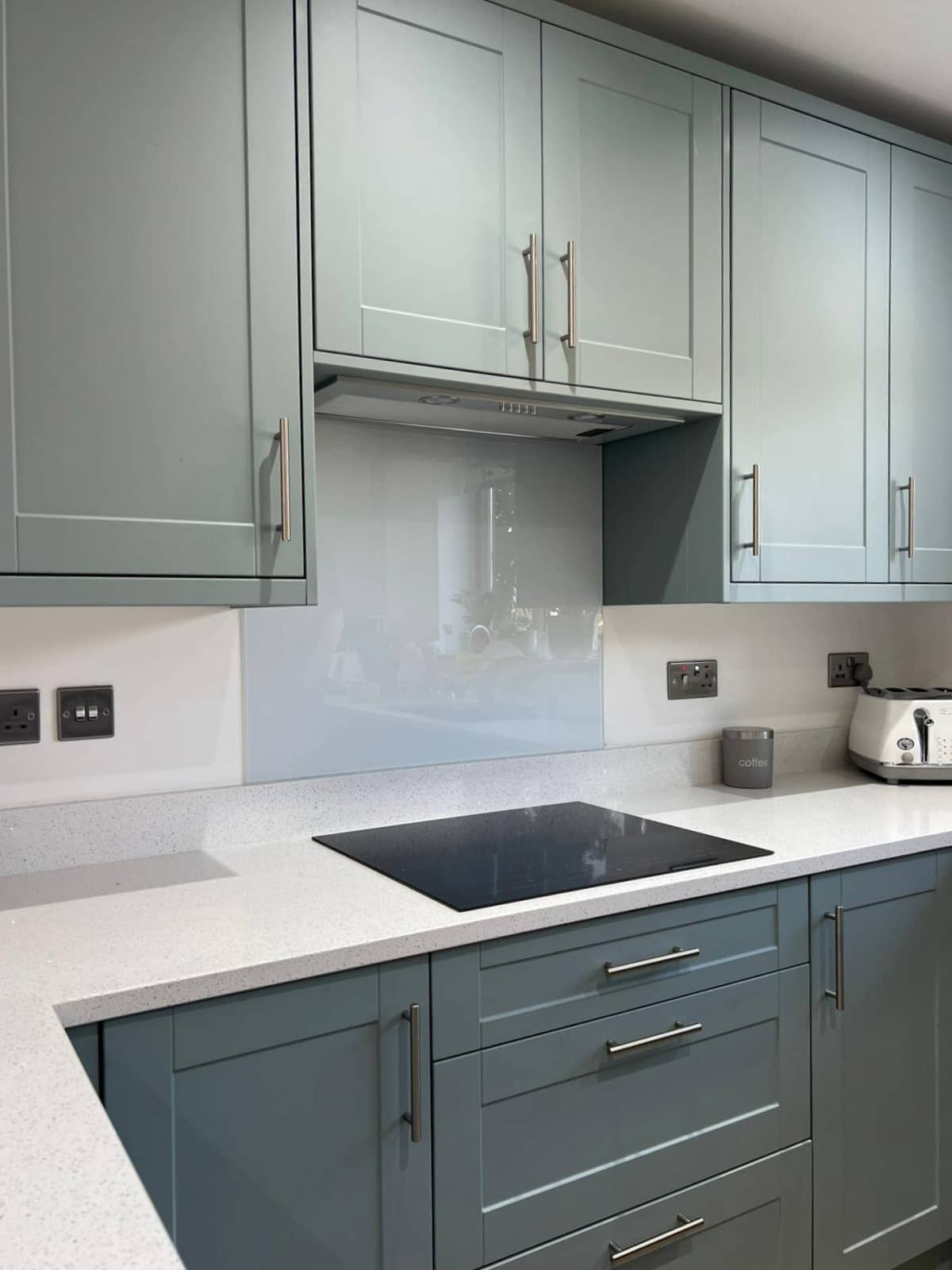 Modern kitchen with light blue cabinets, a white countertop, an electric stovetop, and a light grey backsplash. There are electrical outlets, a white toaster, and a small container labeled "coffee" on the counter.