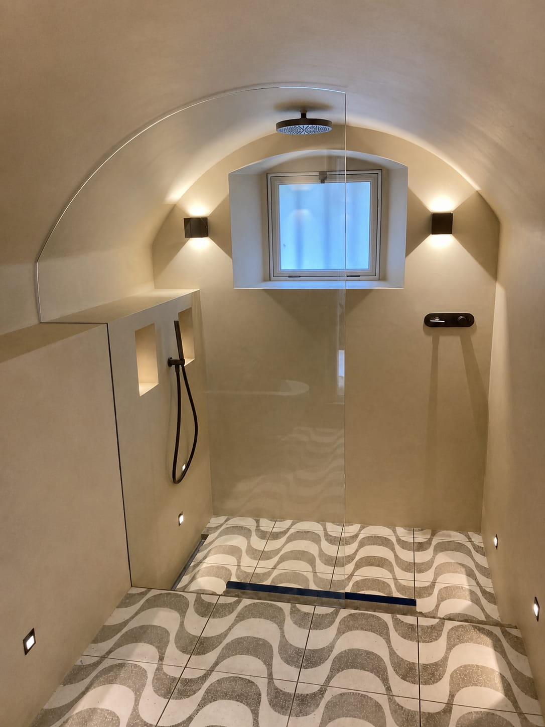 Minimalist beige shower room with a curved ceiling, a rain showerhead, a handheld shower, a frosted window, built-in shelves, black fixtures, and patterned floor tiles with wavy lines.